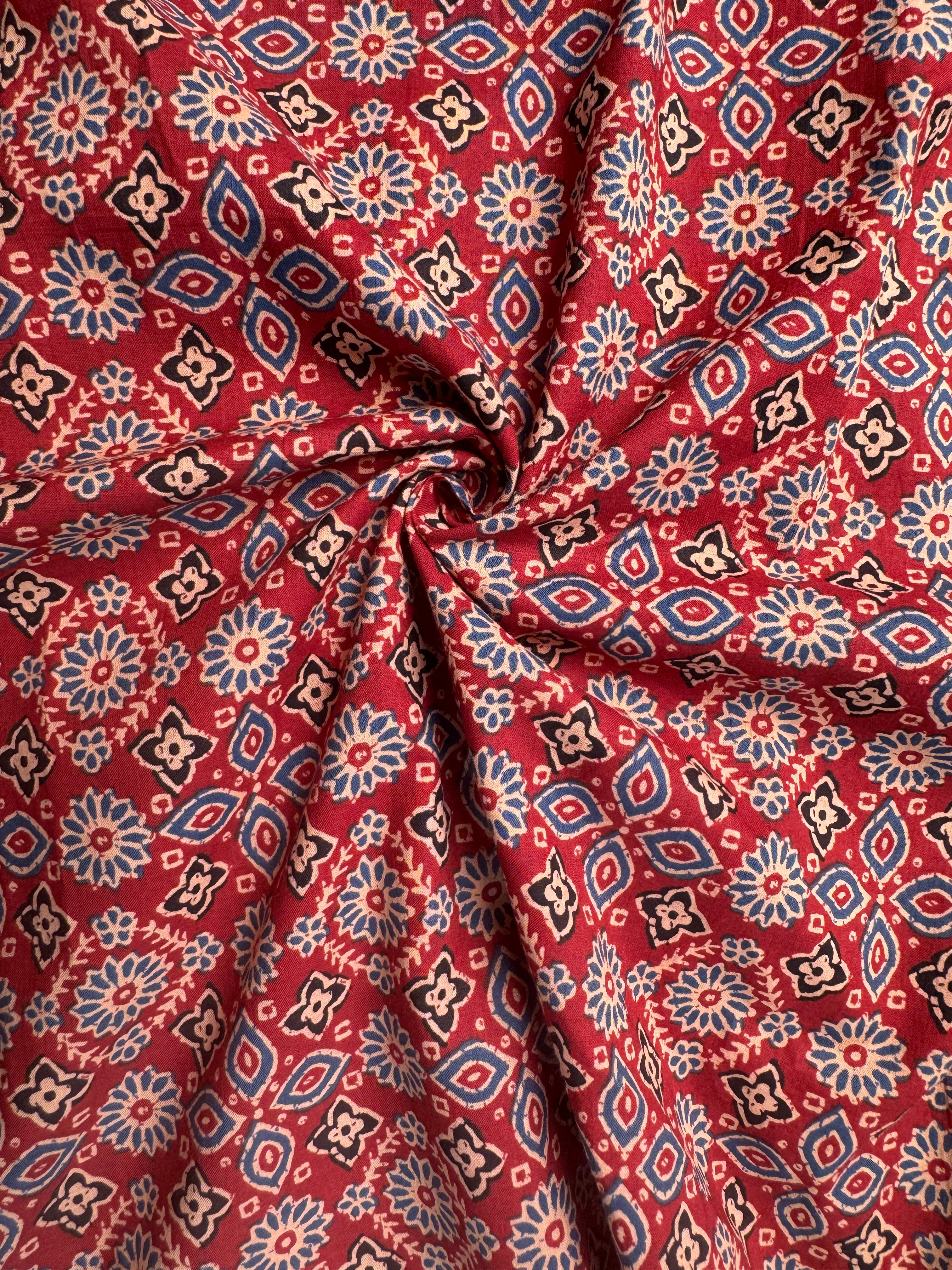Red fabric with a detailed blue, beige, and black geometric and floral pattern, gathered in the center to show its drape and texture.